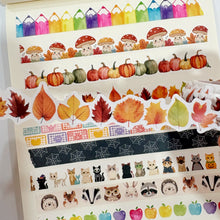 Lade das Bild in den Galerie-Viewer, A collection of decorative washi tapes displayed on a white notebook page. The tapes feature colorful pencil handles, mushrooms, pumpkins, autumn leaves, spiderwebs, cats, raccoons, hedgehogs, and colorful apples.