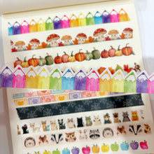 Lade das Bild in den Galerie-Viewer, A close-up of an open notebook or planner showing multiple rows of decorative washi tape. The tapes feature colorful pencils, mushrooms, pumpkins, autumn leaves, cats, owls, raccoons, apples, and vintage-style text patterns.